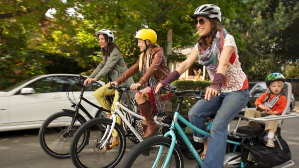 Three women are cycling along an urban road on e-cargo bikes. One has a child on the back of the bike. They are all wearing normal clothes and cycling helmets. There's a white car and trees in the background