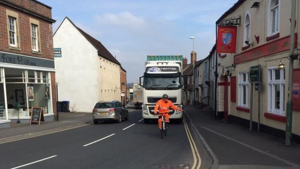 A man in an orange hi-vis outfit is cycling in front of a lorry. He is indicating to go left