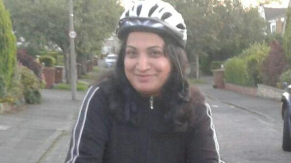 A woman wearing a black jacket and white cycling helmet smiles while holding the handlebars of her bike on a quiet residential street.
