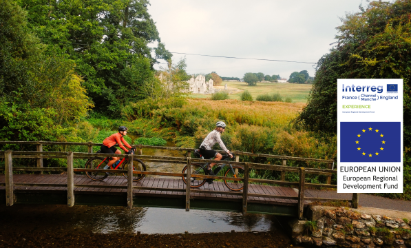 Two people cycle across a wooden bridge with a ruined abbey peeking through the trees behind