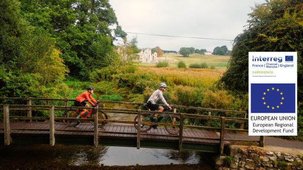 Two people cycle across a wooden bridge with a ruined abbey peeking through the trees behind