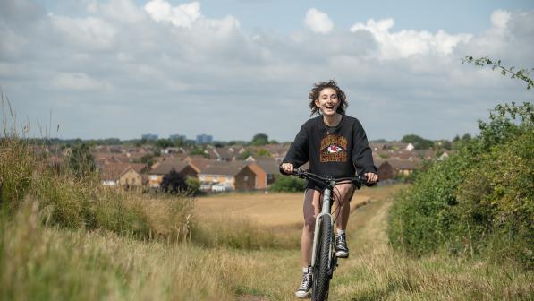 A woman wearing a black jumper and shorts smiles as she pedals an e-cycle uphill through a field. Houses in background.