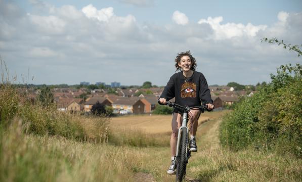 A woman wearing a black jumper and shorts smiles as she pedals an e-cycle uphill through a field. Houses in background.