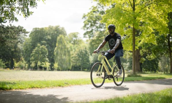A wearing a black t-shirt and helmet pedals an e-cycle through a park on a sunny day