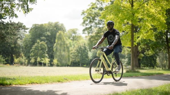 A wearing a black t-shirt and helmet pedals an e-cycle through a park on a sunny day